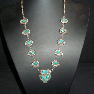 May include: A silver-toned necklace featuring a series of oval-shaped links, each set with a small, smooth turquoise stone. The necklace culminates in a larger, ornate central pendant piece that also contains multiple turquoise stones.