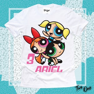 May include: White t-shirt with a graphic of the Powerpuff Girls, Bubbles, Buttercup, and Blossom. The shirt has a pink number 3 and the name "ARIEL" printed on it.