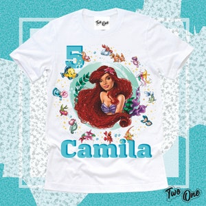 May include: White t-shirt with a colorful illustration of Ariel from The Little Mermaid, surrounded by sea creatures and a number 5. The text "Camila" is printed below the illustration.