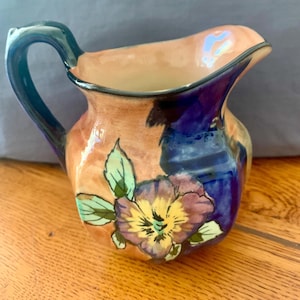 May include: A ceramic jug featuring a glossy finish with a hand-painted pansy flower in shades of purple, yellow, and green. The body of the jug displays a blend of peach and deep blue colours, complemented by a dark blue handle and rim.