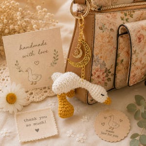 Puede incluir: Un llavero de ganso hecho a mano con ganchillo, con cuerpo blanco y patas y pico amarillos. El llavero cuelga de un dije de media luna dorada. La imagen también muestra un bolso floral, una margarita y tarjetas con texto.