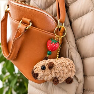 May include: A gold-toned metal keychain featuring a plush, textured light brown capybara charm with dark brown accents, attached to a small green bead and a red strawberry charm with white dots, hanging from a gold-colored metal ring and chain.