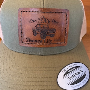 May include: A green and brown trucker hat with a brown leather patch that says "Bronco Life 10/100" and a black and white image of a Ford Bronco. The hat has a white mesh back and a black and white sticker that says "The Authentic Yupoong Snapback Yupoong Since 1974".