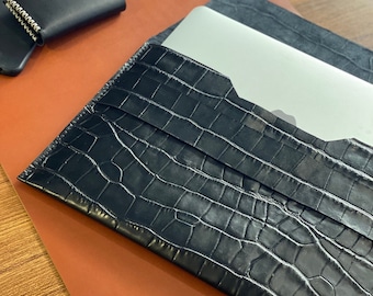May include: Black leather laptop sleeve with a crocodile pattern. The sleeve is closed and lying on a brown leather surface.