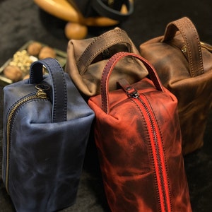 May include: Four leather toiletry bags in different colors: blue, brown, red, and black. The bags have a zipper closure and a leather handle.