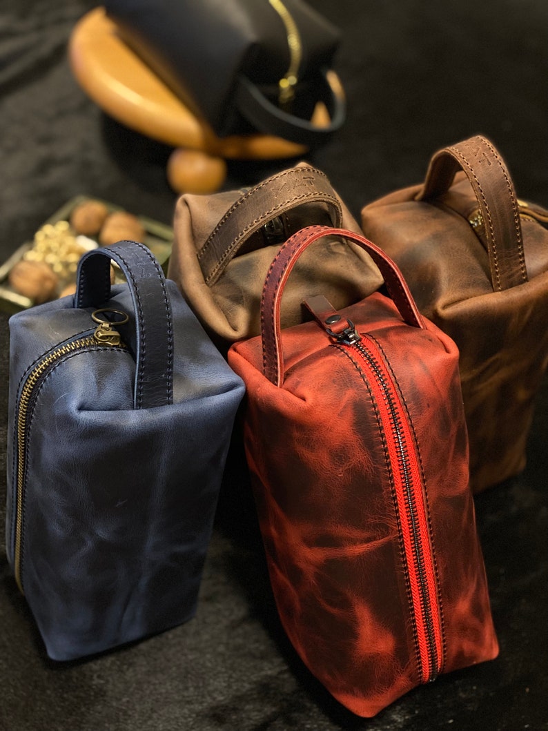 May include: Four leather toiletry bags in different colors: blue, brown, red, and black. The bags have a zipper closure and a leather handle.