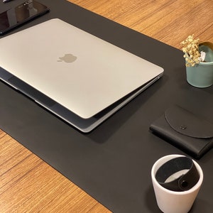 May include: A black leather desk mat with a silver laptop, a black phone, a white pen, a black leather wallet, a white ceramic cup with a black leather band, and a small blue ceramic planter with dried white flowers.