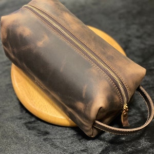 May include: A brown leather toiletry bag with a zipper closure and a brown leather handle. The bag has a distressed look.