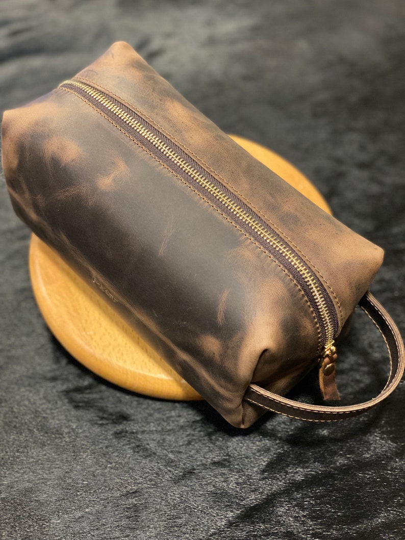 May include: A brown leather toiletry bag with a zipper closure and a brown leather handle. The bag has a distressed look.