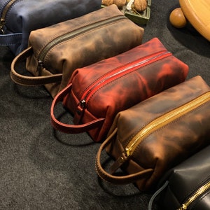May include: Four leather zippered pouches in different colors: blue, brown, red, and black. The pouches have a distressed look and are lined with a gold zipper. The pouches are arranged in a row on a dark gray surface.