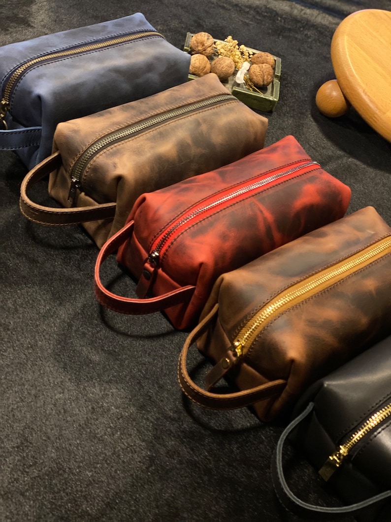 May include: Four leather zippered pouches in different colors: blue, brown, red, and black. The pouches have a distressed look and are lined with a gold zipper. The pouches are arranged in a row on a dark gray surface.