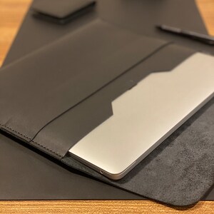 May include: A black leather laptop sleeve with a silver laptop inside. The sleeve is on a black desk mat.