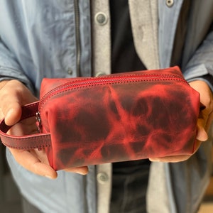 May include: A red leather zippered pouch with a handle. The pouch has a distressed look with a dark red and black color pattern.