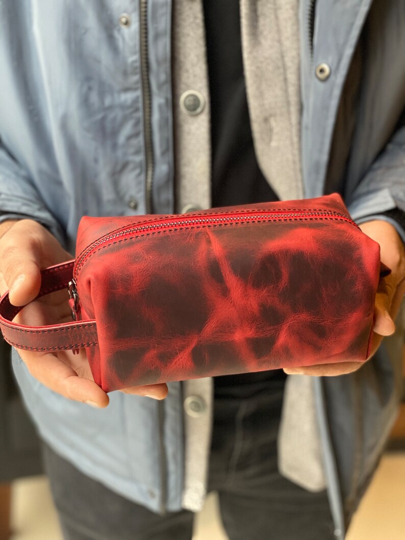 May include: A red leather zippered pouch with a handle. The pouch has a distressed look with a dark red and black color pattern.