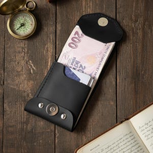 Pode incluir: Uma carteira de couro preta com fecho magnético, contendo notas de lira turca. A carteira está aberta, revelando uma nota de 200 e uma nota de 20 liras turcas. A carteira está sobre uma superfície de madeira.