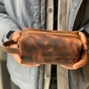 May include: A brown leather zippered pouch with a gold zipper. The pouch has a textured, distressed look.