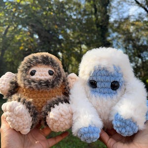 May include: Two handmade crochet plush toys. One is brown and tan, resembling a sasquatch, and the other is white with blue accents, possibly a yeti. Both have black eyes and are made with soft, textured yarn.