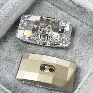 May include: Two rectangular buttons. The top button is clear with a faceted surface and a silver bee design. The bottom button is a muted gold color with a faceted surface and two holes for sewing.