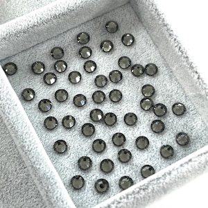 May include: A collection of dark gray, round rhinestones with a faceted surface, scattered inside a light gray velvet tray. The rhinestones are evenly spaced and reflect light, creating a sparkling effect. Ideal for crafting or jewelry making.