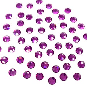 May include: A scattering of small, round, faceted, magenta-colored rhinestones. Each gem has a flat back and a multi-faceted top, catching the light. The rhinestones are scattered across a white background, creating a sparkling effect.