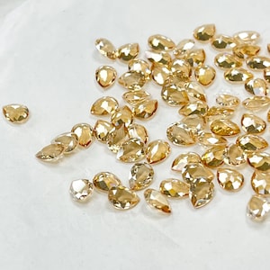 May include: A collection of numerous small, teardrop-shaped golden crystals with multifaceted surfaces. Each individual crystal reflects light, creating a shimmering effect across the group. The pieces are scattered in a loose arrangement.