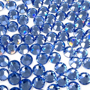 May include: A collection of round, faceted, sapphire-blue rhinestones. The gemstones are scattered across a white surface, each catching the light and displaying a spectrum of colors. These sparkling embellishments are ideal for craft projects.