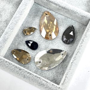 May include: Assortment of teardrop-shaped rhinestones in various colors, including clear, black, and shades of gold and brown. The rhinestones are displayed in a gray velvet tray, showcasing their faceted surfaces and reflective qualities.