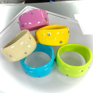 May include: A collection of five colorful bangles in shades of pink, yellow, light blue, green, and cream. Each bangle is adorned with small, sparkling rhinestones. The bracelets are made of a glossy material.