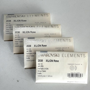 May include: Four packages of Swarovski Elements crystals are stacked. Each package is labeled with the Swarovski Elements logo, the crystal type, and size. The packages are labeled "2038 XILION Rose" and include details like "SS 34", "SS 30", "SS 20", and "SS 16".