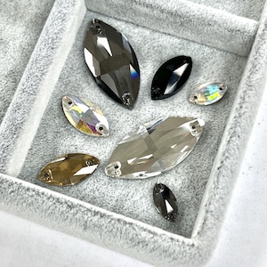 May include: A collection of marquise-shaped rhinestones in various colors, including clear, black, and iridescent, are arranged in a gray velvet tray. The rhinestones have two holes for sewing or attaching. The tray is a soft gray color.
