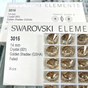 May include: A close-up of a package of Swarovski Elements crystals. The package includes the text "SWAROVSKI ELEMENTS" and "3015 14 mm Crystal (001) Golden Shadow (GSHA)". The crystals are a golden colour and are arranged in a grid.