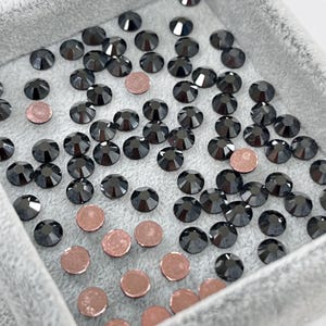 May include: A collection of black, faceted rhinestones and rose gold flat-backed embellishments are scattered inside a gray, fabric-lined tray. The rhinestones have a glossy finish, and the rose gold pieces are circular.