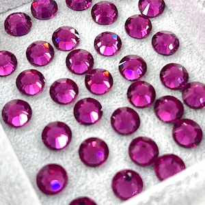 May include: Close-up of numerous round, faceted, magenta-colored rhinestones. The gems are scattered on a light gray surface, each reflecting light. The rhinestones are a vibrant shade of pink, perfect for crafting or embellishment.