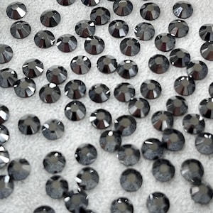 May include: A collection of dark gray, faceted rhinestones scattered on a white, textured surface. Each rhinestone has a circular shape with multiple facets, creating a sparkling effect. The rhinestones are uniform in size and shape.