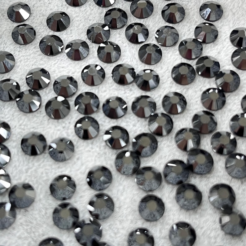 May include: A collection of dark gray, faceted rhinestones scattered on a white, textured surface. Each rhinestone has a circular shape with multiple facets, creating a sparkling effect. The rhinestones are uniform in size and shape.