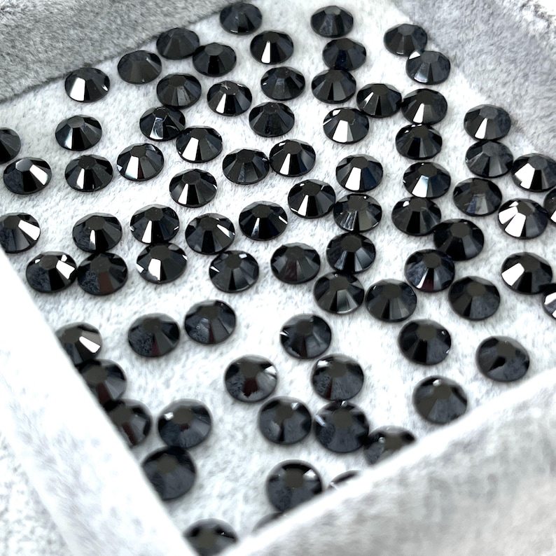 May include: A collection of black, faceted rhinestones, arranged in a gray velvet-lined tray. The rhinestones are round with a flat back and a multi-faceted top, reflecting light. The tray is partially visible, providing a soft backdrop for the sparkling stones.