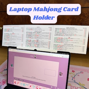 May include: A white laptop Mahjong card holder with printed text. The holder is positioned above a laptop displaying a Mahjong game interface. The text on the holder includes "Laptop Mahjong Card Holder" and game-related information.