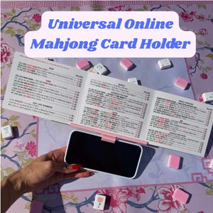 May include: A light pink plastic smartphone stand holding a white Mahjong card with printed game rules and point values. The stand is positioned on a floral patterned surface, surrounded by scattered Mahjong tiles with pink and white backs.