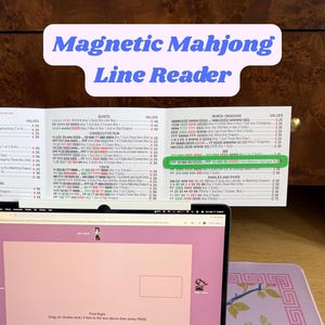 Puede incluir: Una pantalla de ordenador muestra un gráfico "Magnetic Mahjong Line Reader". El gráfico enumera varias combinaciones, valores y categorías "Winds-Dragons", con texto verde resaltado. Una computadora portátil y una superficie rosa están en primer plano.