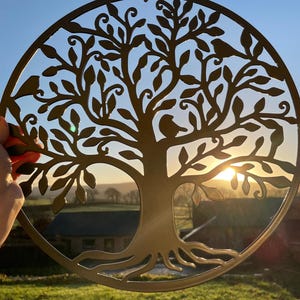 May include: A gold metal tree of life wall decoration with a circular design and intricate details. The tree has a thick trunk and branches with leaves and birds. The background is a blurry image of a field and a house.