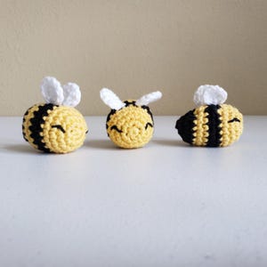 May include: Three crocheted bumblebees with black and yellow stripes and white wings. The bumblebees are smiling and have black eyes.
