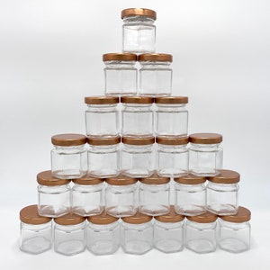 May include: A pyramid arrangement of twenty-one small, clear glass jars with hexagonal bases and metallic copper-colored screw-on lids. Each jar is empty and features a transparent body, creating a uniform display of containers stacked in rows.