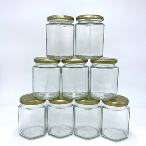 May include: A stack of nine clear glass jars with hexagonal bases and gold-colored lids. The jars are empty and arranged in a pyramid shape against a white background. The jars are suitable for food storage or crafting.