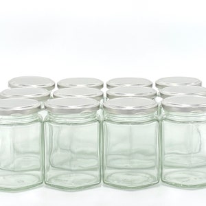 May include: A dozen clear glass jars with silver lids are arranged in a grid. The jars have a hexagonal base and a rectangular body. The lids are flat and metallic. The jars are empty and sit against a white background.