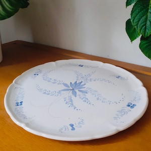 May include: A white ceramic serving plate with a blue floral design. The plate has a scalloped edge and is decorated with a stylized flower and leaves.