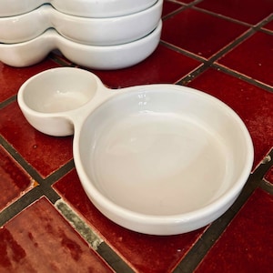 Puede incluir: Un plato de cerámica blanca para doble inmersión con dos cuencos redondos conectados por un asa. El plato está sobre una superficie de azulejos rojos.