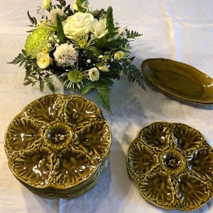 Peut inclure: Un ensemble de six assiettes à huîtres en céramique verte, avec une pile de quatre assiettes au centre et deux assiettes individuelles de chaque côté. Les assiettes ont un motif en relief de coquilles d'huîtres.