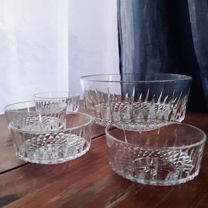 Puede incluir: Conjunto de cinco cuencos de vidrio transparente con un patrón de diamante. Los cuencos están dispuestos sobre una superficie de madera. El cuenco más grande está en la parte posterior y el cuenco más pequeño está en la parte delantera.