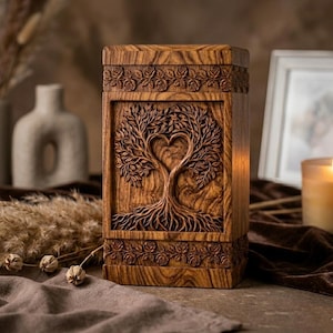 May include: A wooden urn featuring a carved tree design, with branches forming a heart. The rectangular urn has floral carvings at the top and bottom. The wood has a warm, natural tone. A candle and decorative items are in the background.