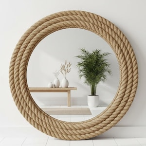 Pode incluir: Espelho redondo com uma moldura de corda espessa e texturizada. A corda bege natural cria uma estética náutica ou costeira. O espelho reflete uma sala com uma parede branca, um banco de madeira e uma planta em vaso, criando uma sensação brilhante e arejada.